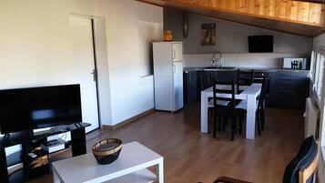 Ein kompakter Wohnraum mit Fernseher, Esstisch, Stühlen und einer Küchenzeile mit Kühlschrank sowie einem Fenster.