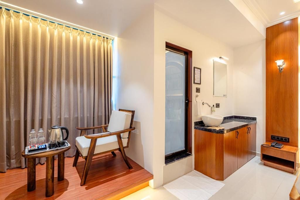 Quarto de hotel com uma cadeira de madeira, uma mesinha com uma cafeteira, uma pia com espelho e um armário de madeira.