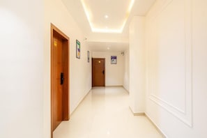 Un pasillo con una puerta de madera a la izquierda, una pared blanca con una puerta al final e iluminación empotrada en el techo.