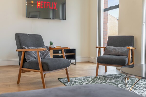 A modern living room with two grey armchairs, a small side table with a plant, and a TV mounted on the wall displaying 'NETFLIX'. - Luxury 4 Bed | Jewellery Quarter | Private Parking (Birmingham)