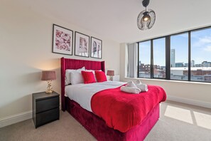 Habitación de hotel con una cama grande, una mesita de noche, una lámpara y vistas de la ciudad a través de un ventanal amplio.