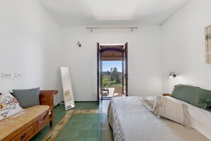 Ein Schlafzimmer mit einem Bett, einer hölzernen Bank, einem kleinen Tisch und Blick durch eine offene Tür auf eine Terrassenanlage.