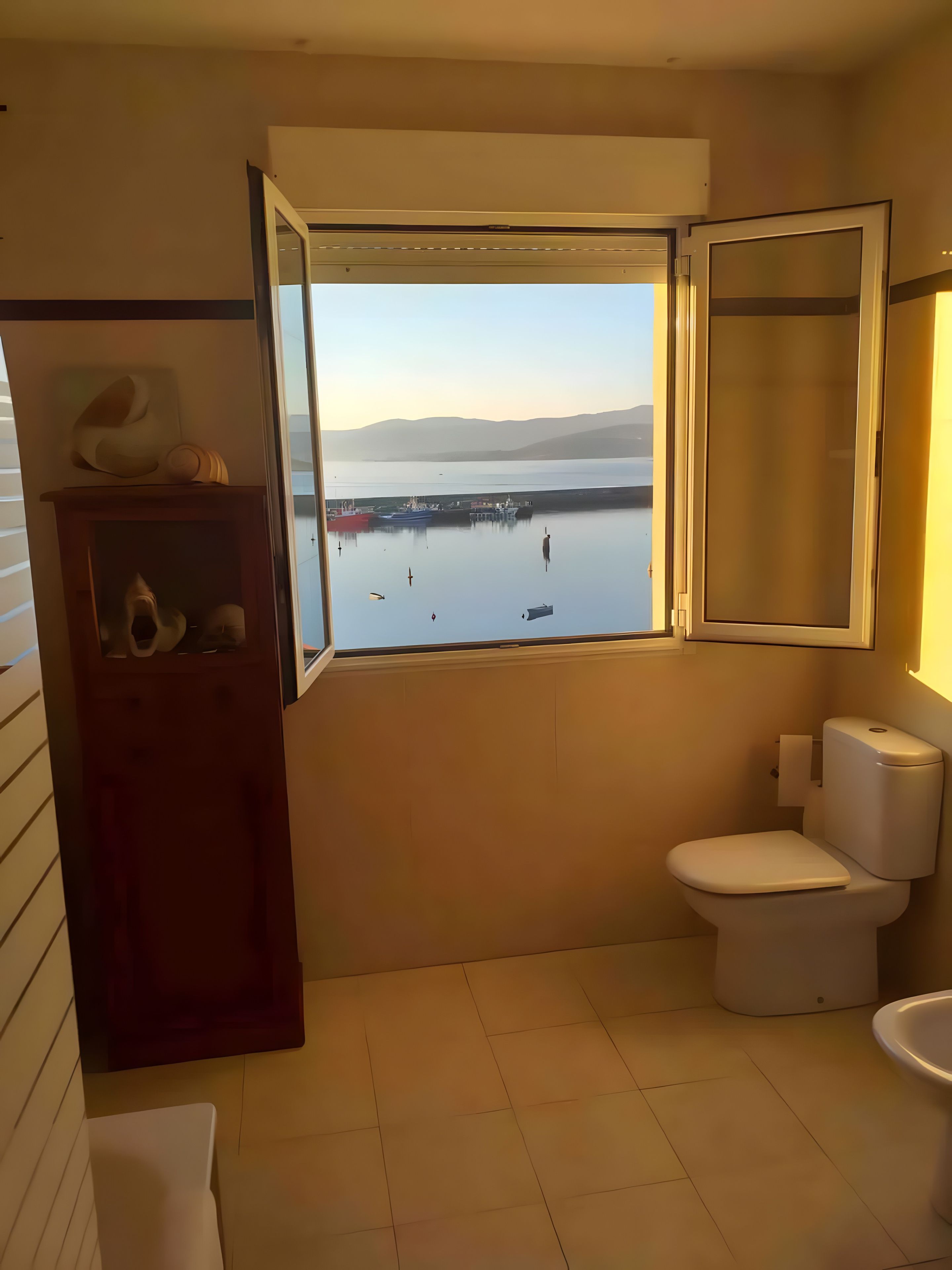 Un baño con inodoro, lavamanos, ventana con vistas al agua y barcos, y un armario de madera con zapatos.