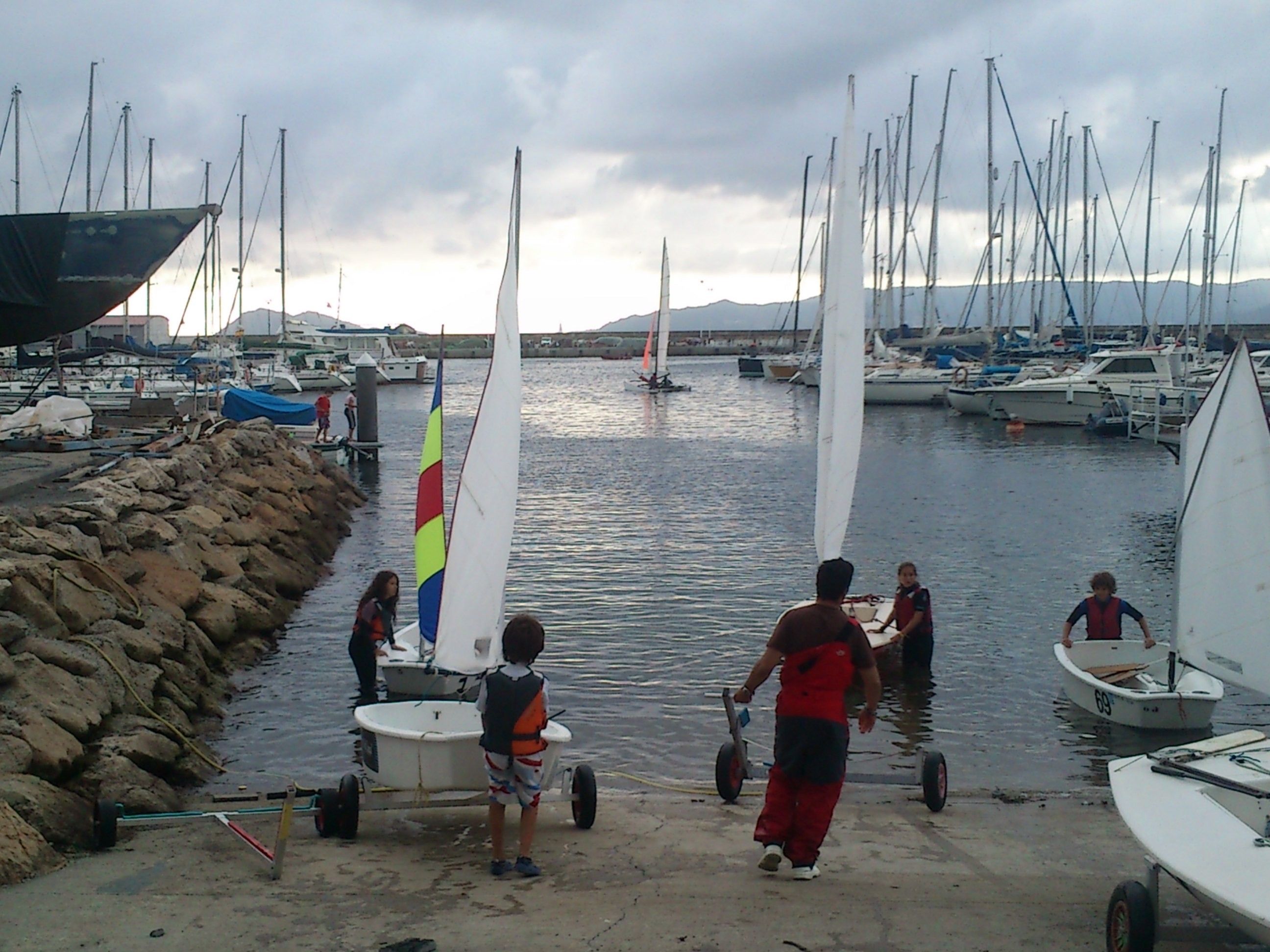 A marina with multiple sailboats docked, a sandy shore, and a rocky breakwater.