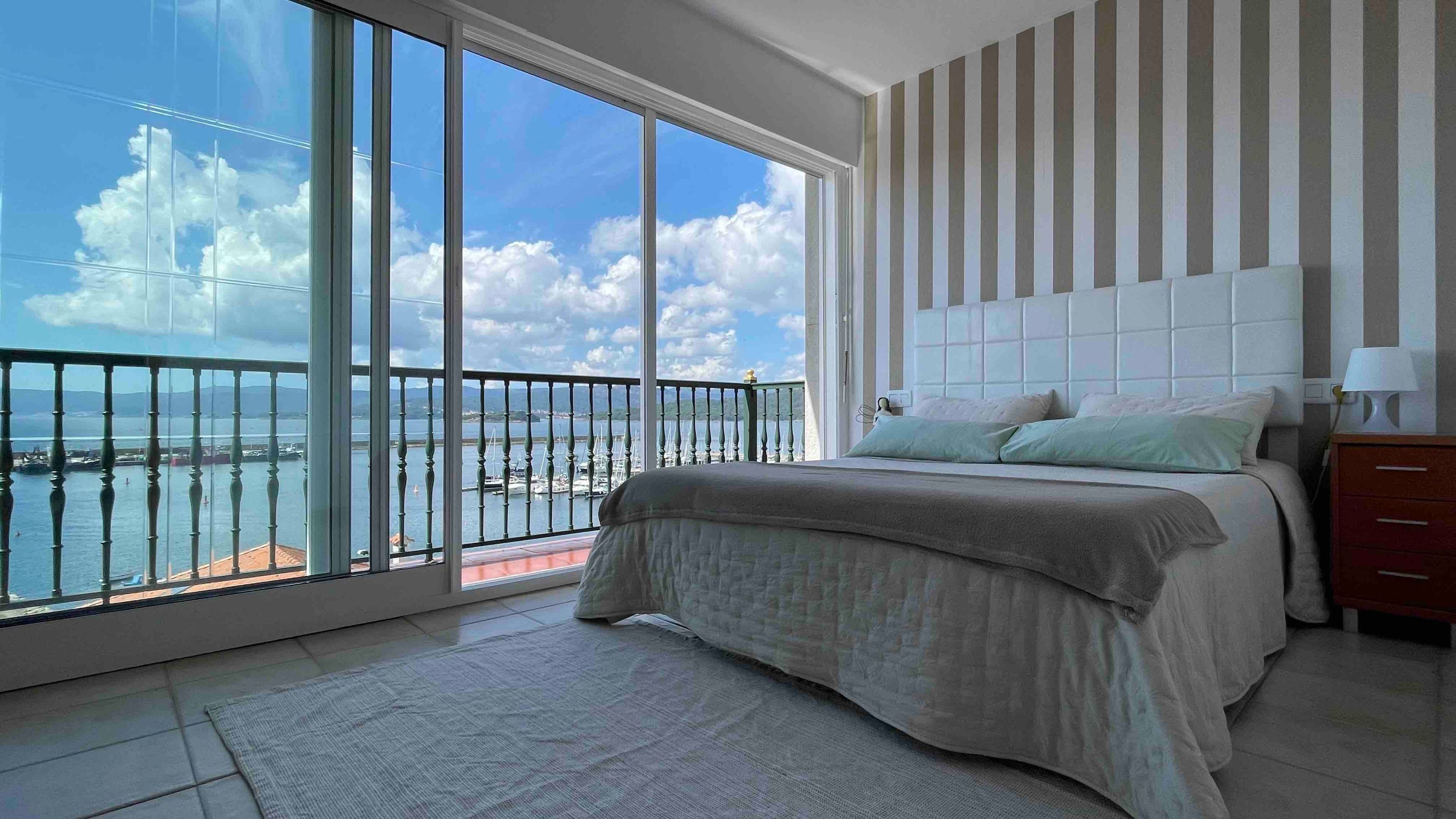 A bedroom with a large window, a bed, a nightstand, and a view of a body of water.