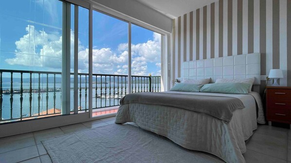 A bedroom with a large window, a bed, a nightstand, and a view of a body of water.