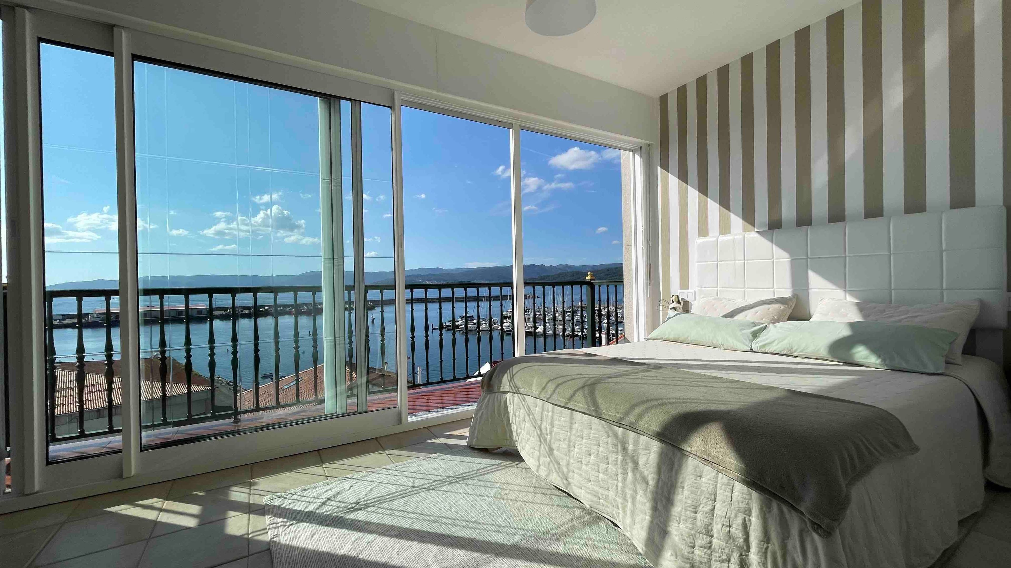 A bedroom with a large sliding glass door leading to a balcony with a view of the sea and boats.