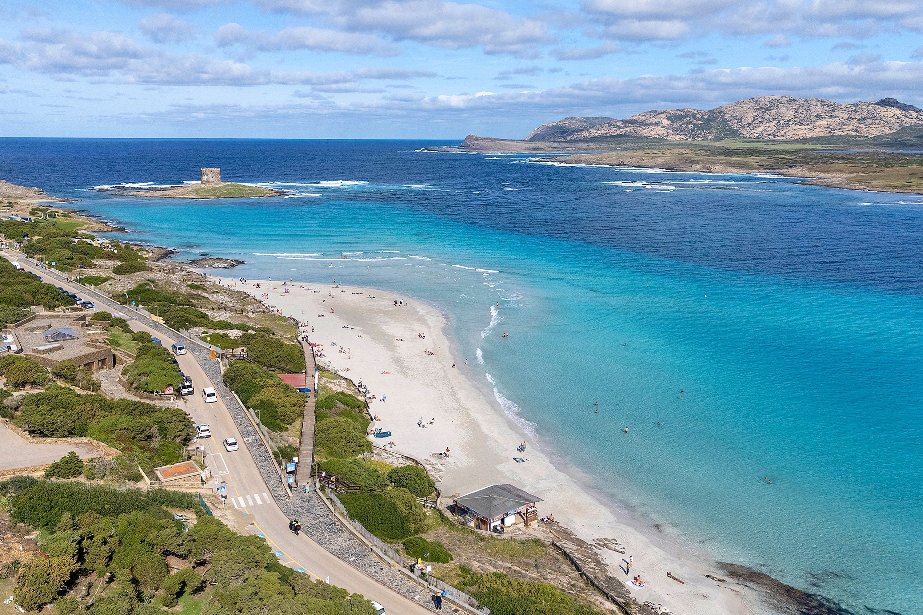 Une route côtière qui mène à une plage aux eaux d’un bleu limpide, avec une petite tour et, en arrière-plan, des collines rocheuses.