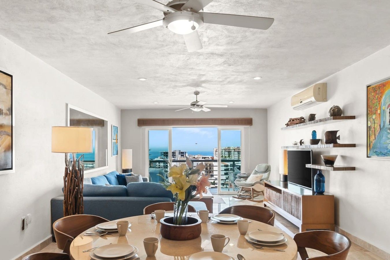 A modern living room with a dining area, a ceiling fan, and a view of the city.