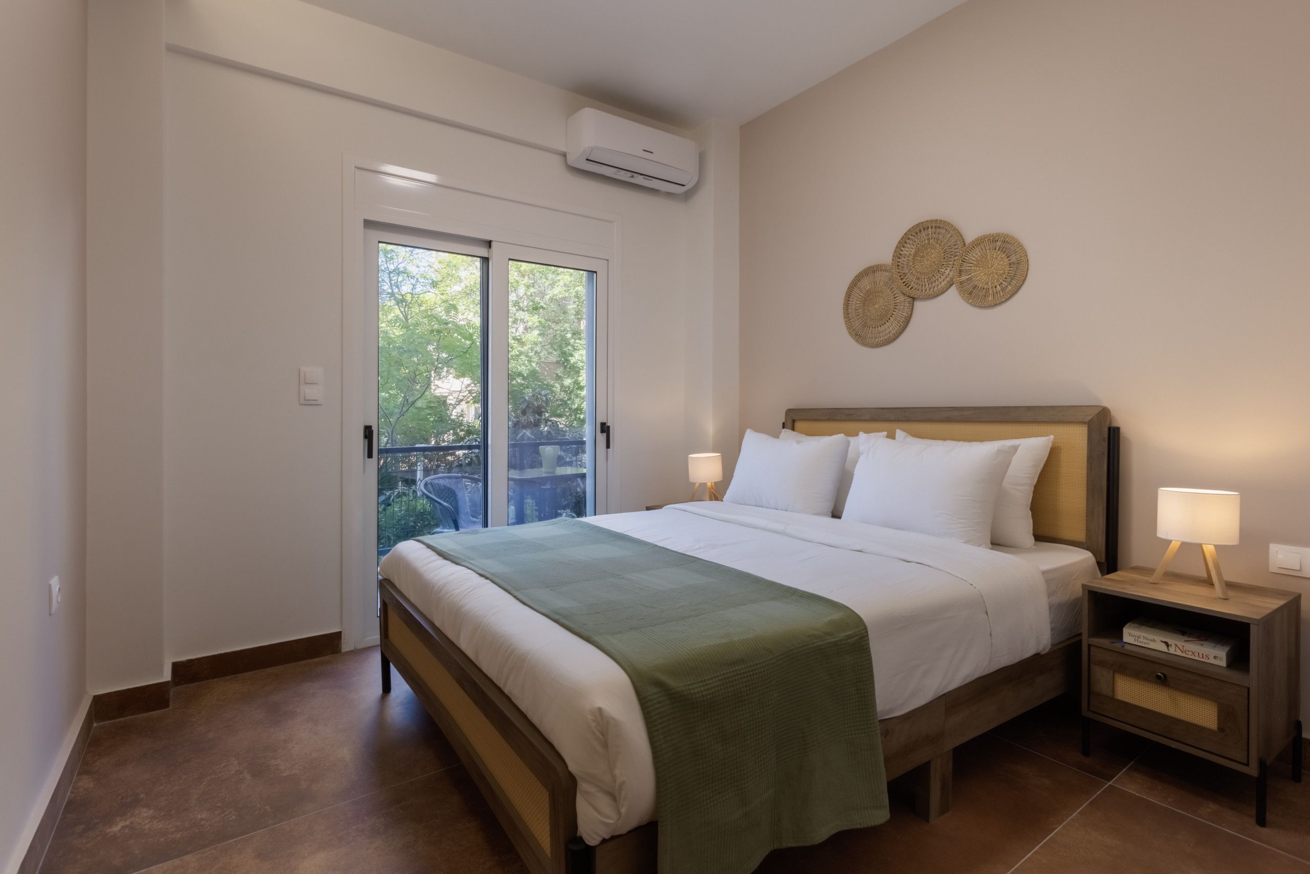 A bedroom with a bed, bedside lamps, a nightstand, a wall-mounted air conditioner, and a view through a glass door.