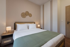 A neatly made bed with white linens and a green throw blanket, flanked by two bedside tables with lamps, in a room with a wooden wardrobe and a wall-mounted decorative item. - Harokopou 3·HM1·ground (Kallithea)