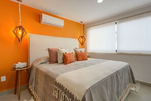 A bedroom with a bed, pillows, a nightstand, an air conditioning unit, and a window with blinds. - Cozy Modern Condo Steps to Everything (Puerto Vallarta)