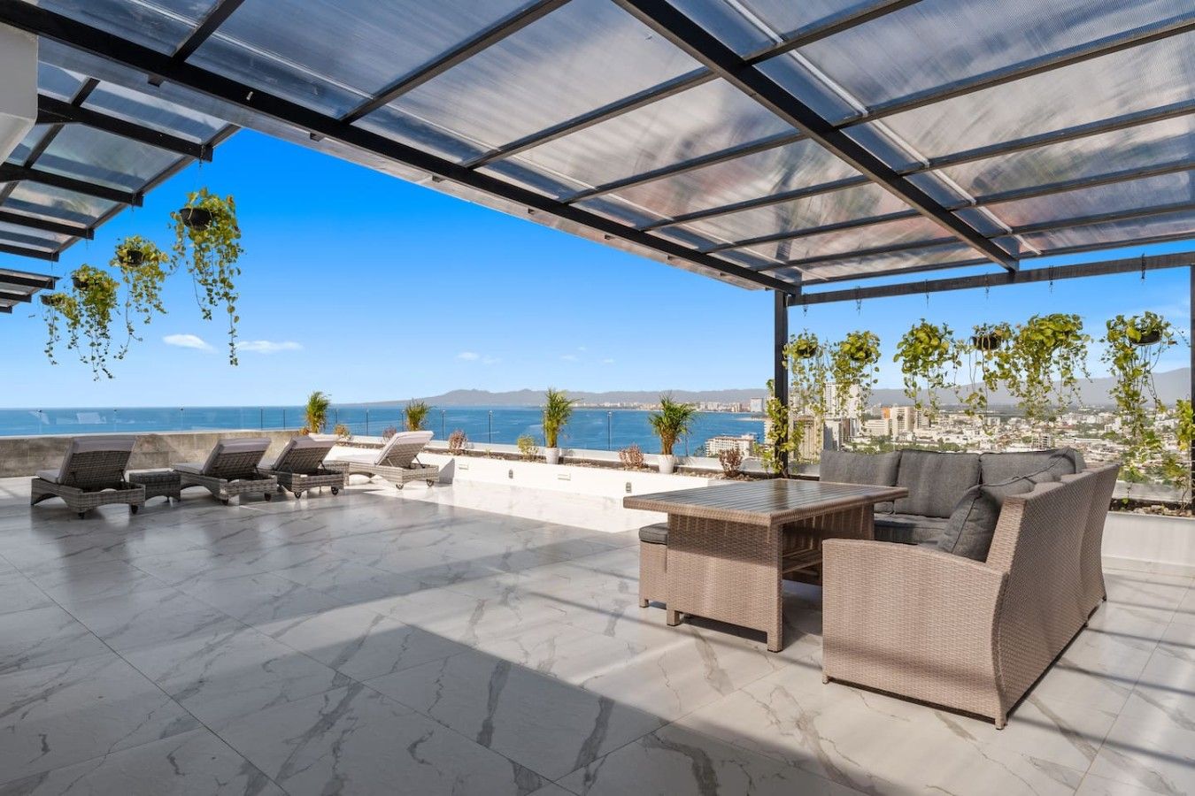 An outdoor patio with a view of the sea, furnished with wicker furniture, and hanging plants.