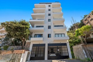 Um edifício de vários andares com entrada central, varandas em cada pavimento e vegetação ao redor.