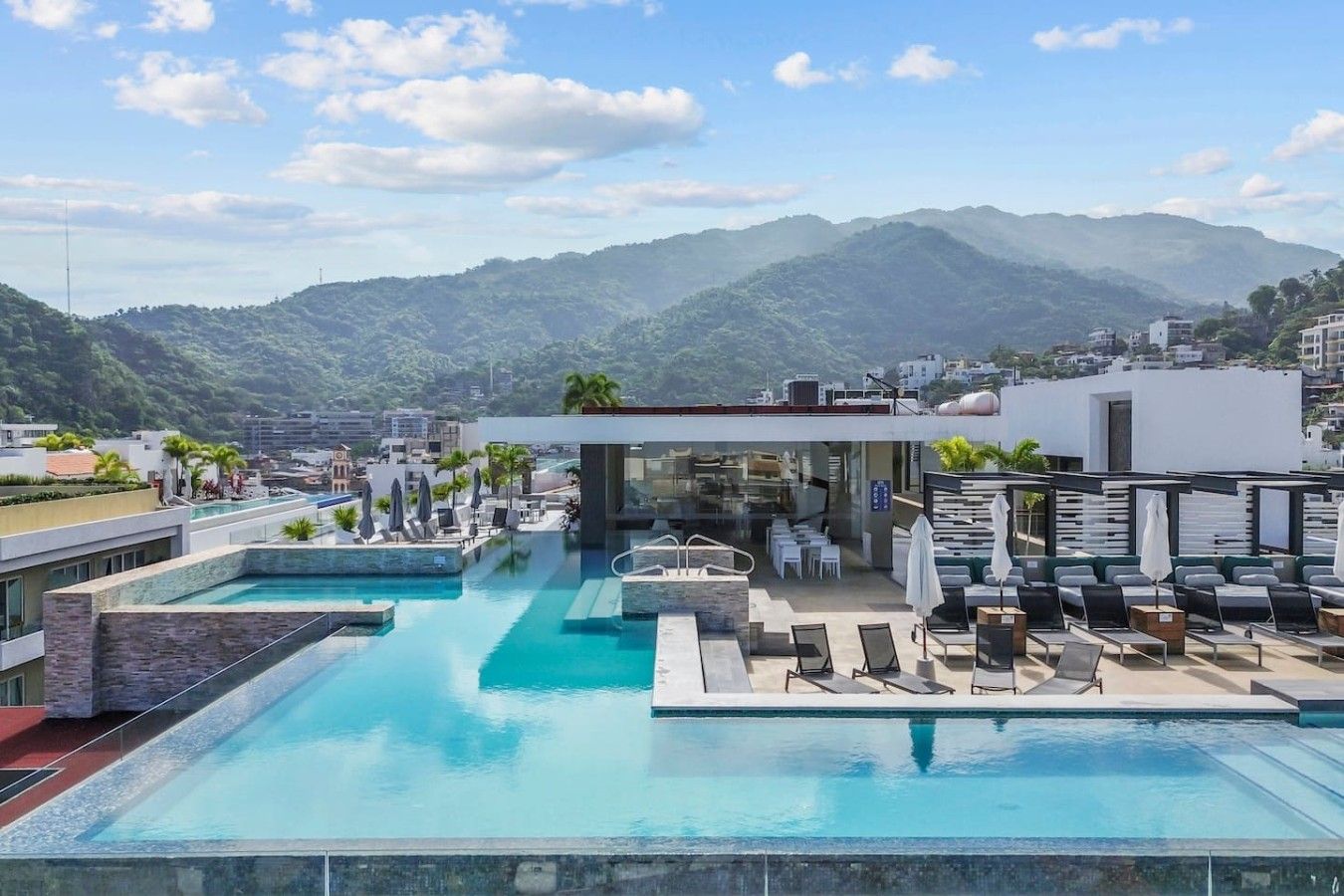 A rooftop pool area with multiple swimming pools, lounge chairs, and a restaurant.