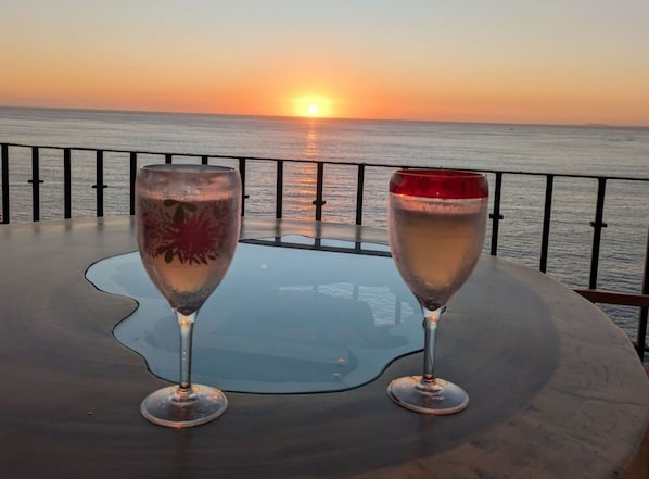 Deux verres d’une boisson sont posés sur une table, offrant une vue sur le coucher de soleil sur l’océan.