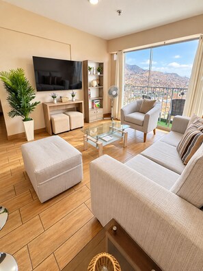 A modern living room with a flat-screen TV, comfortable seating, a coffee table, and a view of a cityscape through large windows.
