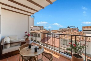 Ein Balkon mit einer Couch, einem runden Tisch und Stühlen mit Blick auf eine Stadtlandschaft und terrakottafarbene Dächer.