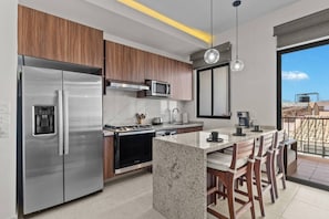 Eine moderne Küche mit einem Edelstahl-Kühlschrank, einem Einbauofen und einer Mikrowelle. Es gibt eine große Kücheninsel mit Barhocker und einen Blick auf die Stadt durch ein großes Fenster.