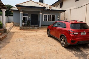 A residential property with a paved driveway, a house with a tiled roof, and a red SUV parked in the driveway. - Serene 1BR 1BT Wi-Fi Kitchen Laundry TV Wardrobe (Ejisu)