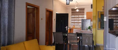 A living room with a yellow sofa, a dining area with chairs and a table, and a kitchen in the background.