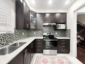 A modern kitchen with dark cabinetry, a stainless steel stove, and a tiled backsplash. - Bright Home: Furnished Patio, Fast WiFi & Parking (Brampton)
