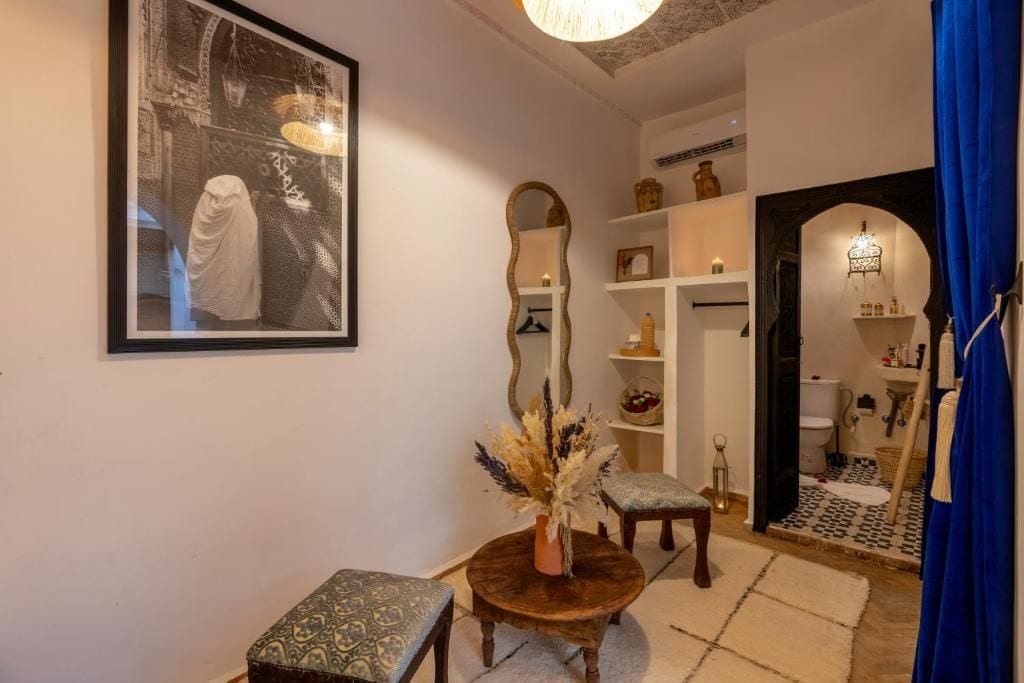 A room with a large framed photograph, a round wooden table with a vase, a patterned bench, and a mirror on the wall.