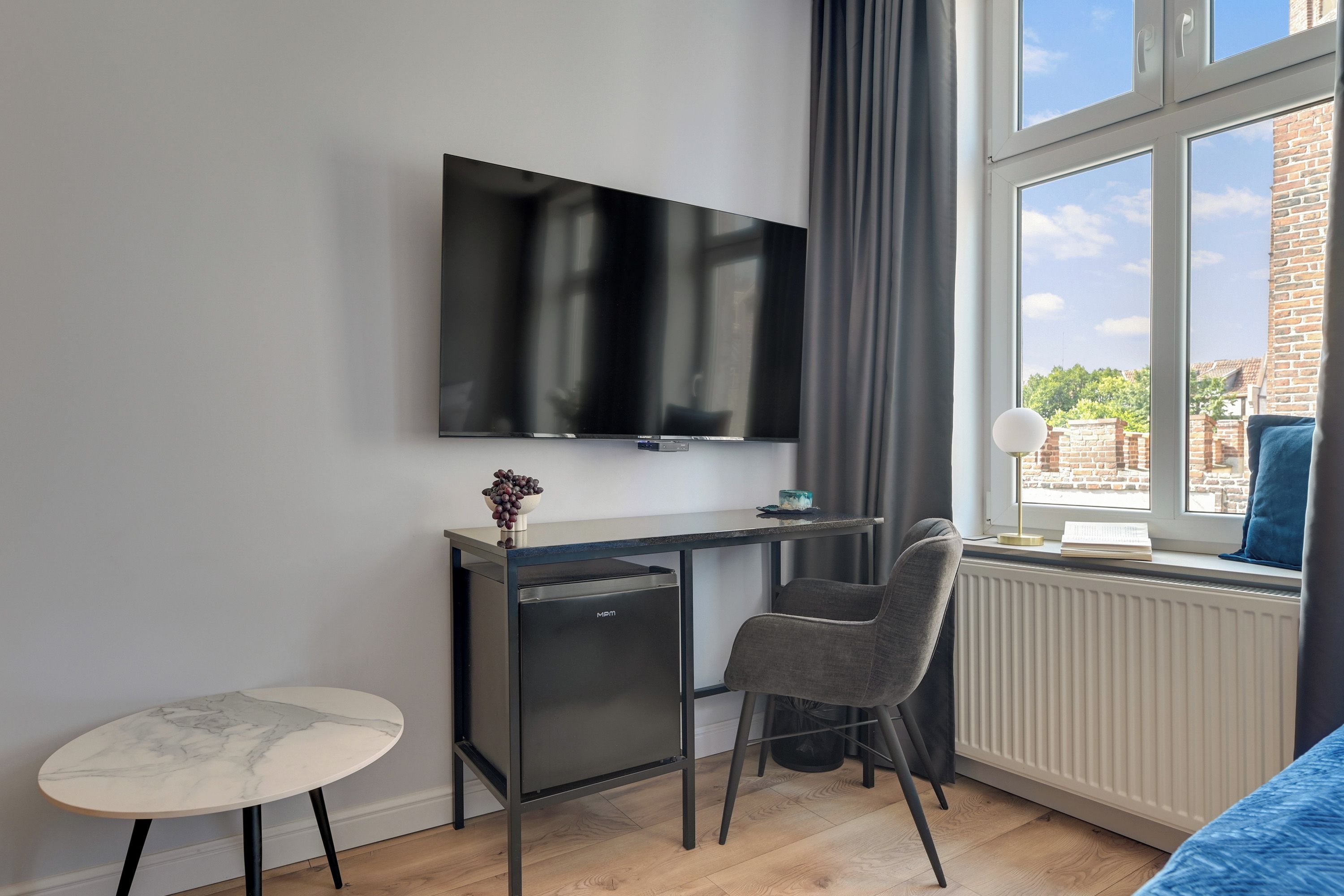 Una habitación moderna con un televisor montado en la pared, un escritorio pequeño con una silla, una mesa redonda y una ventana con vistas a edificios.