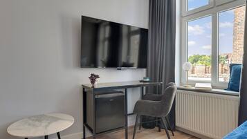 Una habitación moderna con un televisor montado en la pared, un escritorio pequeño con una silla, una mesa redonda y una ventana con vistas a edificios.