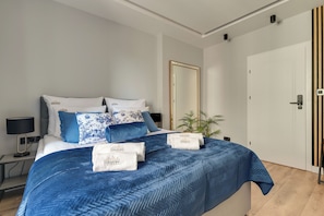 Quarto de hotel moderno com uma cama grande, mesa de cabeceira, luminária, espelho e porta com painel de madeira vertical.