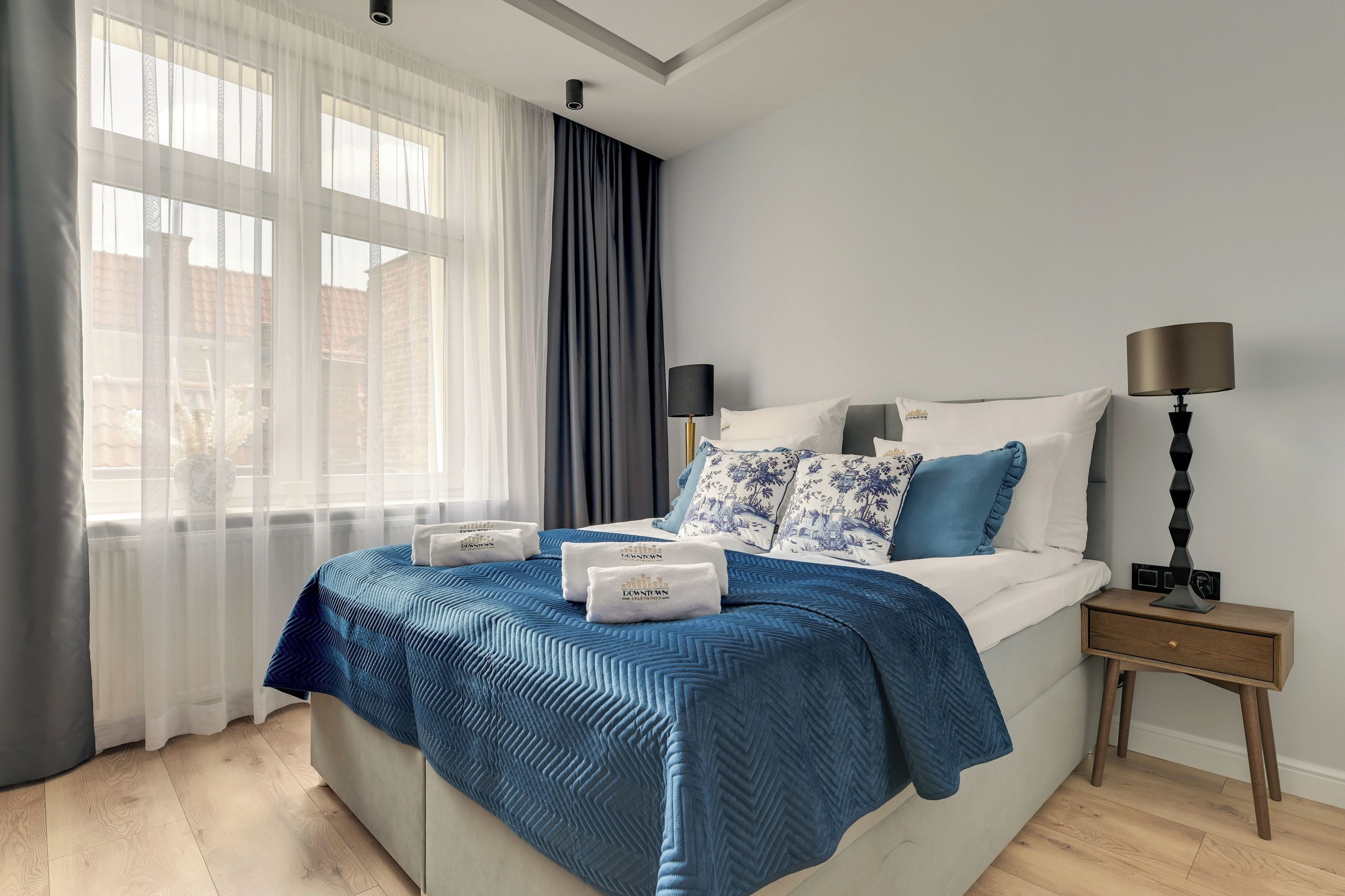 Una habitación de hotel con cama, mesita de noche, lámpara y ventana con cortinas.