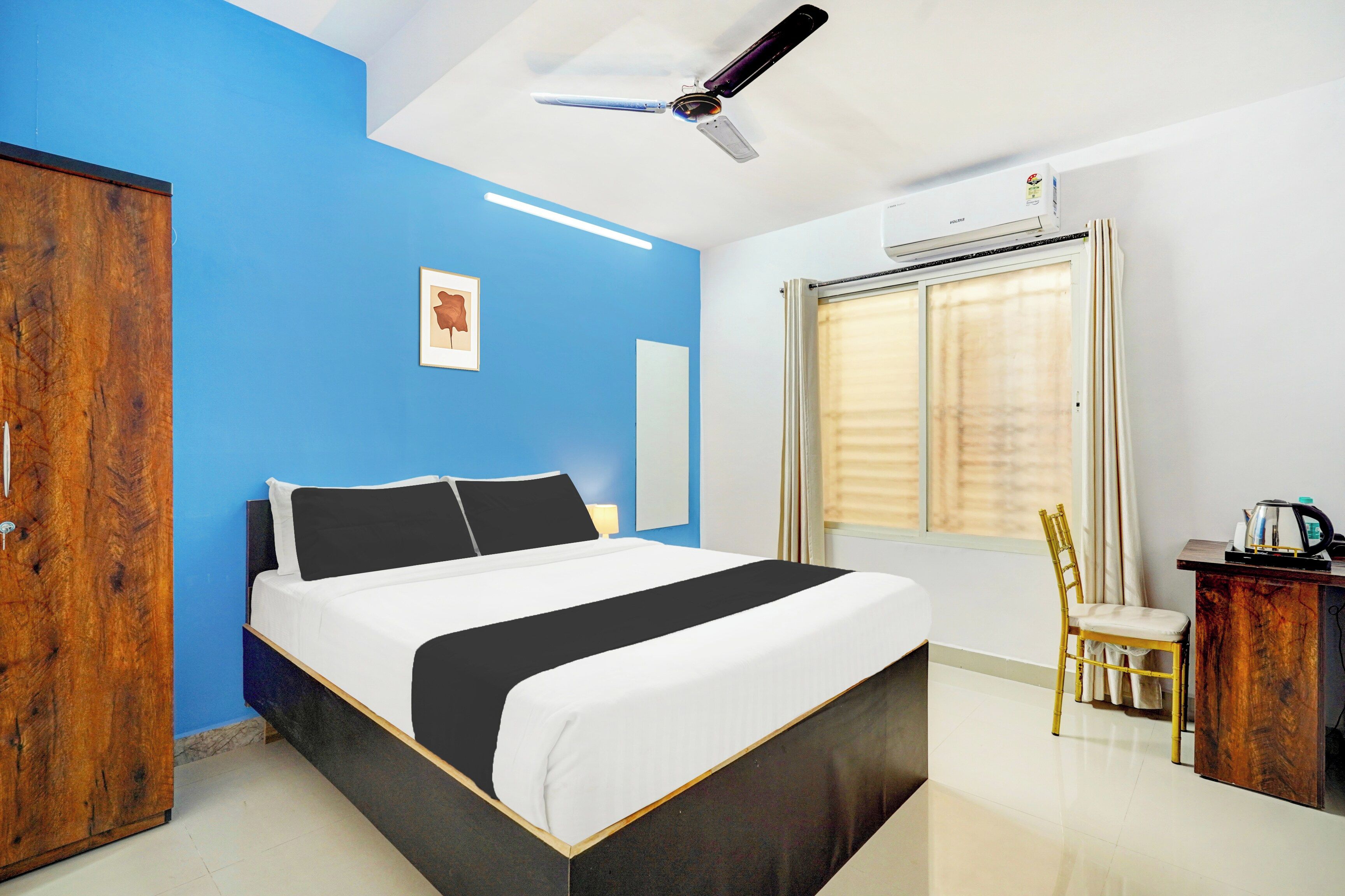 Une chambre d’hôtel comprenant un lit, un bureau, une chaise, une armoire, un ventilateur de plafond et une fenêtre avec des stores.