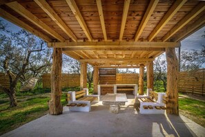 Une structure de pergola en bois, dotée de bancs et d’une table centrale, entourée d’un jardin.