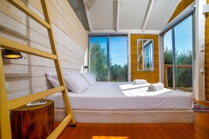 Ein Schlafzimmer mit einem hölzernen Bett, einer Himmelsleiter, einer Fernbedienung und Blick auf Bäume durch große Fenster.