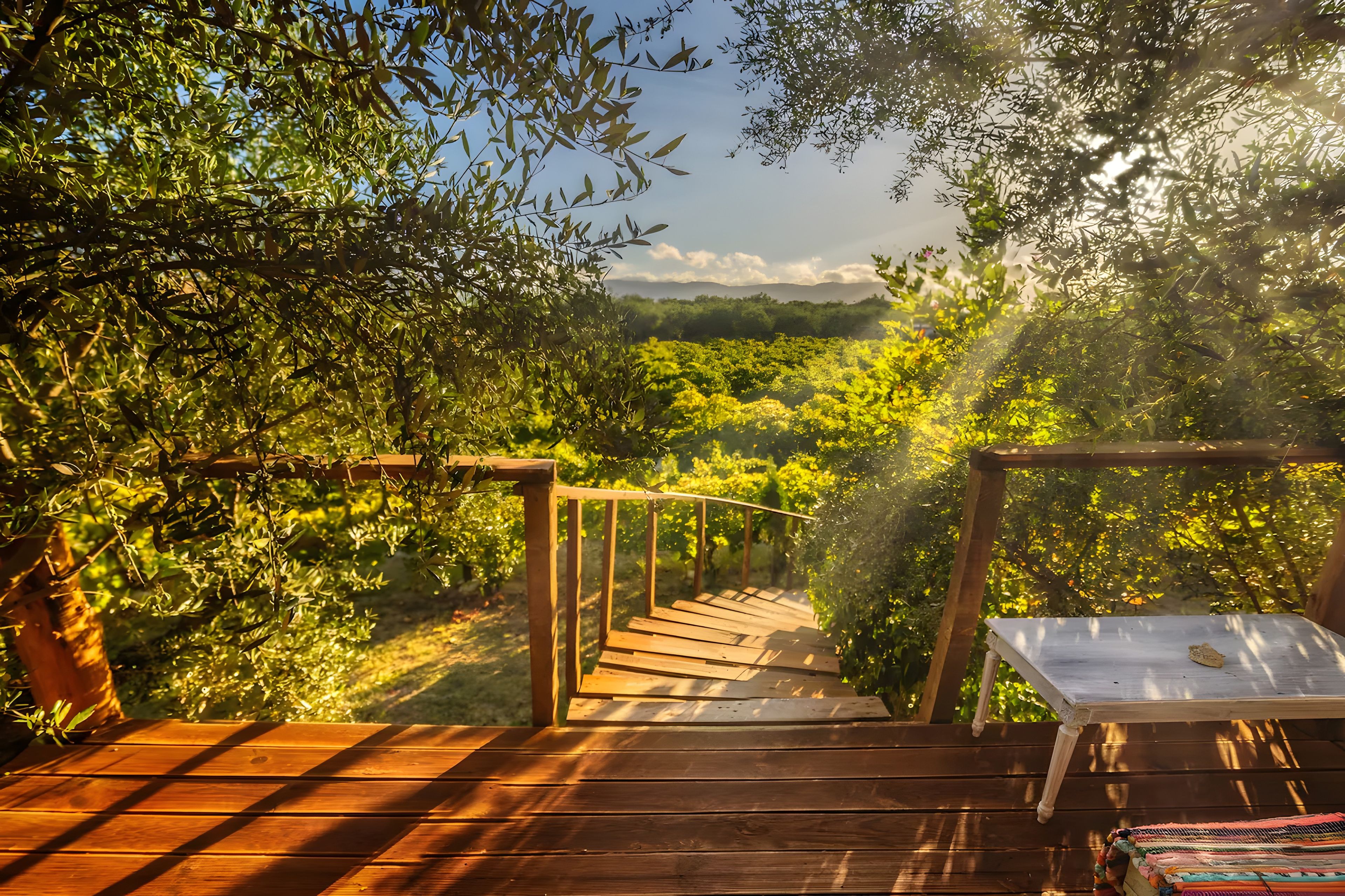 Una terraza de madera que lleva a un espacio cubierto con una mesa y sillas, rodeado de exuberante vegetación y vistas a colinas lejanas.