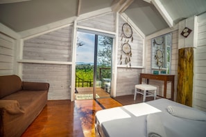 A room with a wooden floor, a bed, a sofa, a desk, and a view through a glass door to an outdoor area with a wooden deck and greenery. - Ferienhaus 'Terra Vine Collection' (Αρκάδιοι)