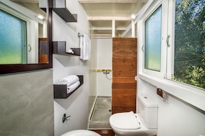 A compact bathroom with a toilet, a wooden shower enclosure, and a window with a view of trees. - Ferienhaus 'Terra Vine Collection' (Αρκάδιοι)