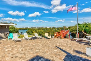 A patio with lounge chairs, a flagpole with an American flag, and a view of a body of water and buildings in the distance. - Fishing Pier | Heated Community Pool | Quick Walk to the Beach | Sea Shell #32 (Indian Shores)