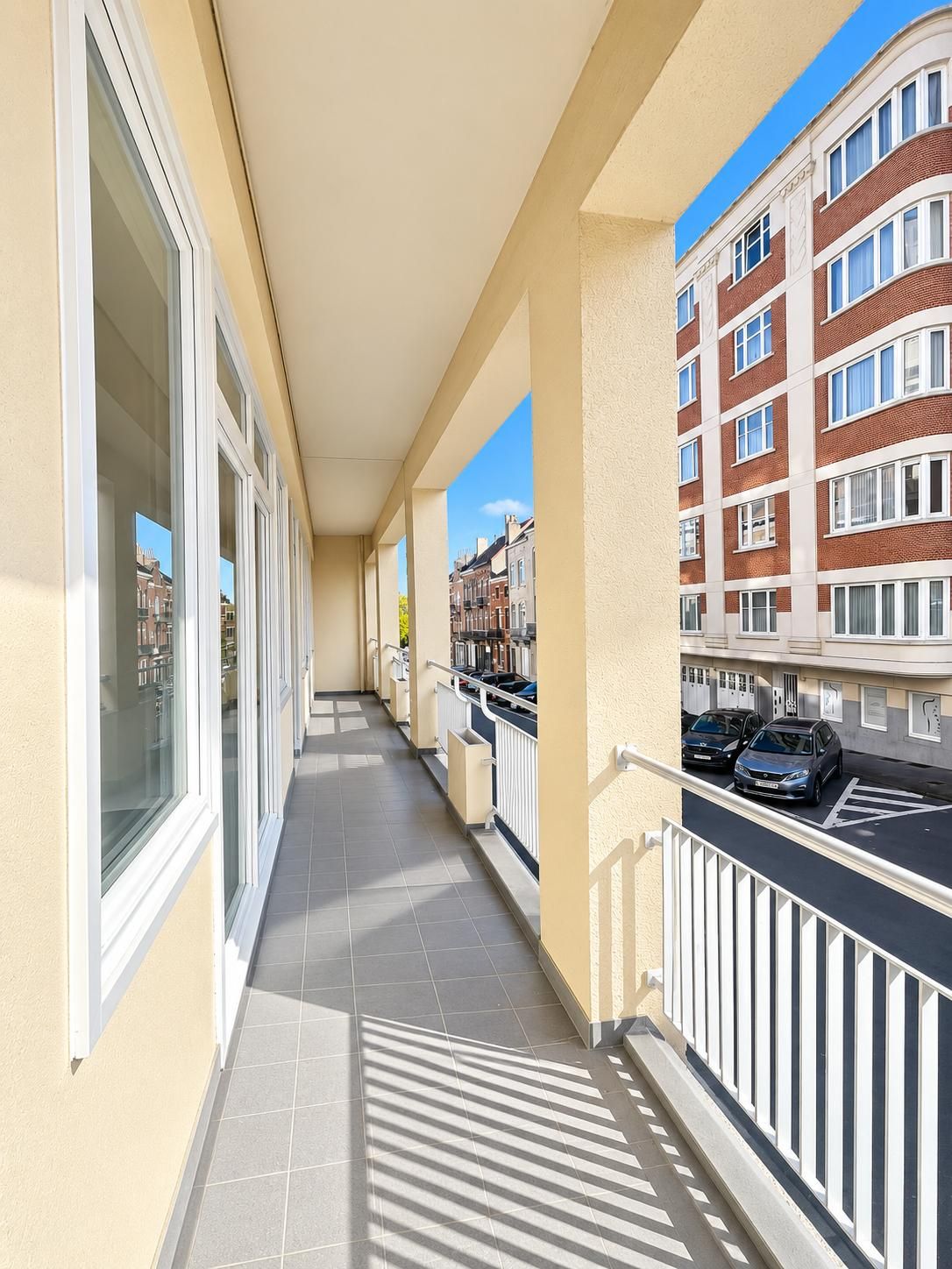 A corridor with a tiled floor, a balcony with a railing, and a view of a multi-story building with windows.