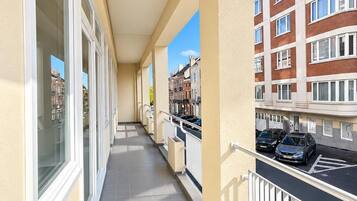 A corridor with a tiled floor, a balcony with a railing, and a view of a multi-story building with windows.