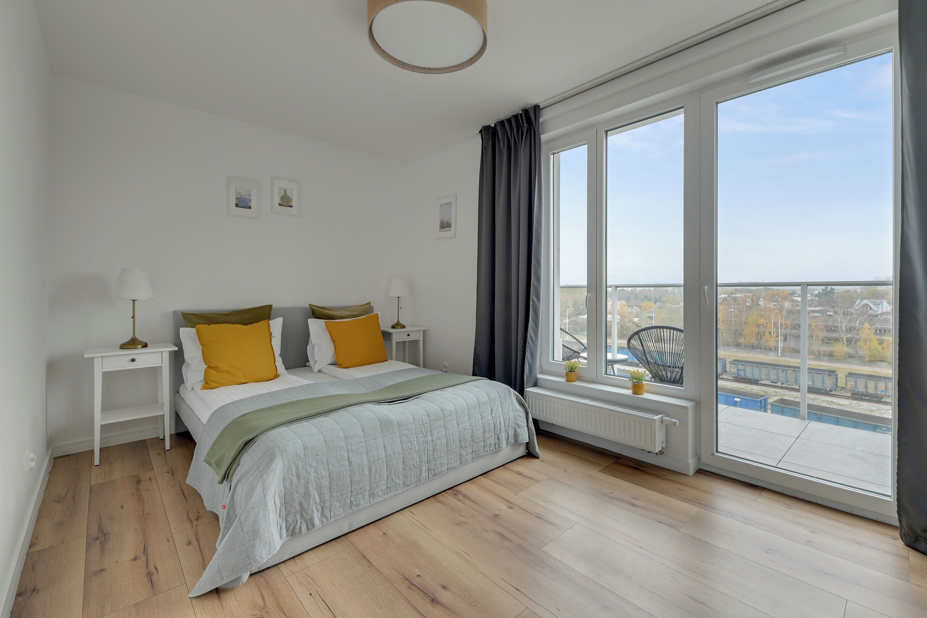 A bedroom with a large window, a bed with a grey duvet, two bedside tables with lamps, and a view of a cityscape.