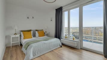 A bedroom with a large window, a bed with a grey duvet, two bedside tables with lamps, and a view of a cityscape.