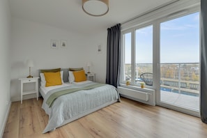 Un dormitorio con un ventanal grande, una cama con cubrecama gris, dos mesitas de noche con lámparas y vistas a un paisaje urbano.