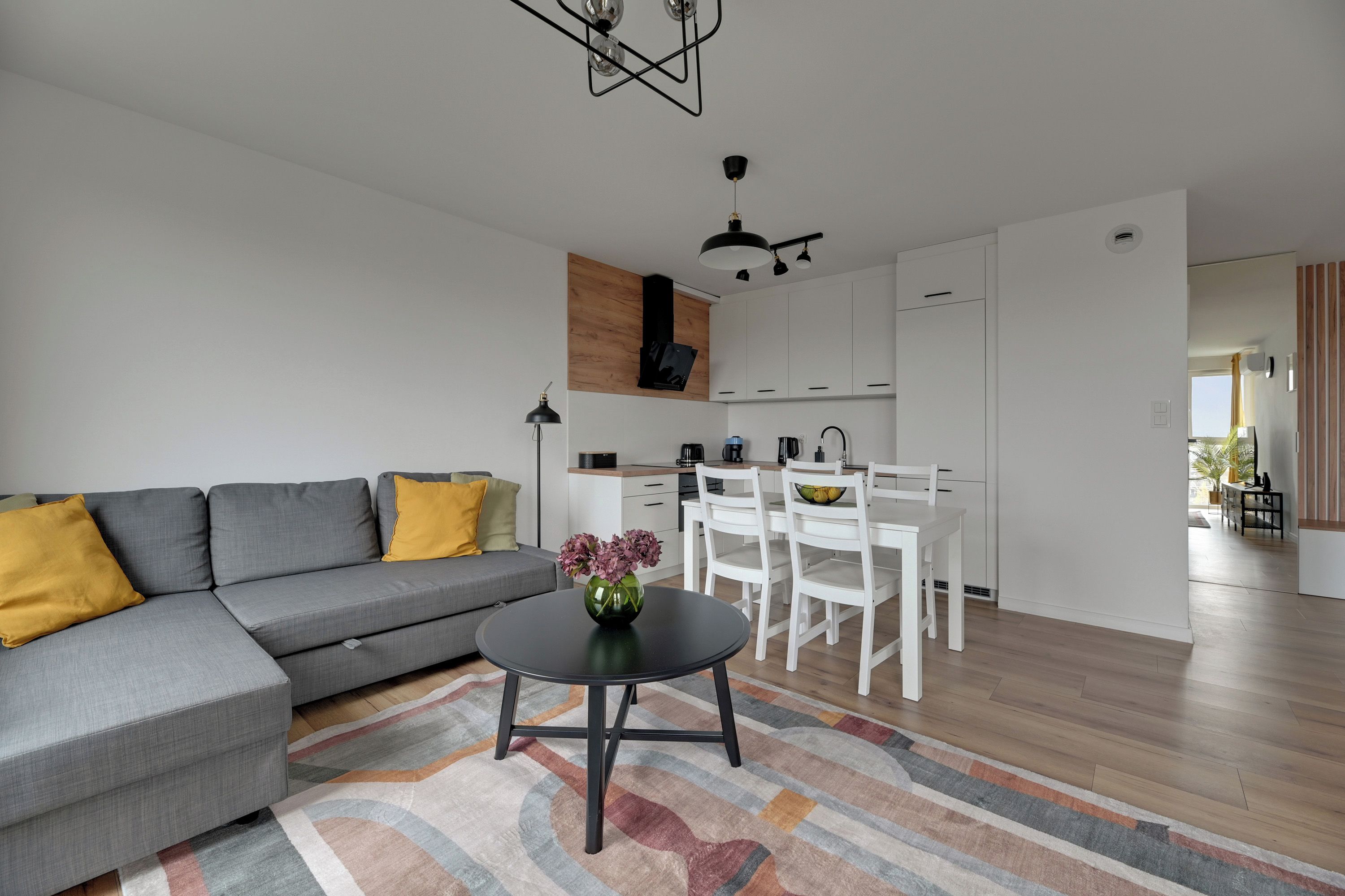 Un salon moderne avec un canapé d’angle gris, une petite table ronde ornée d’un vase et un tapis aux couleurs vives.