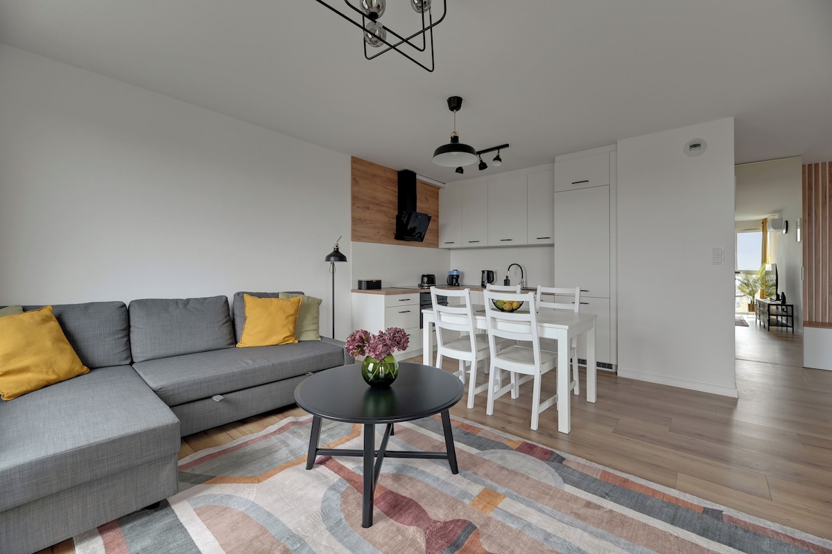 A modern living room with a grey sectional sofa, a small round table with a vase, and a colorful rug.