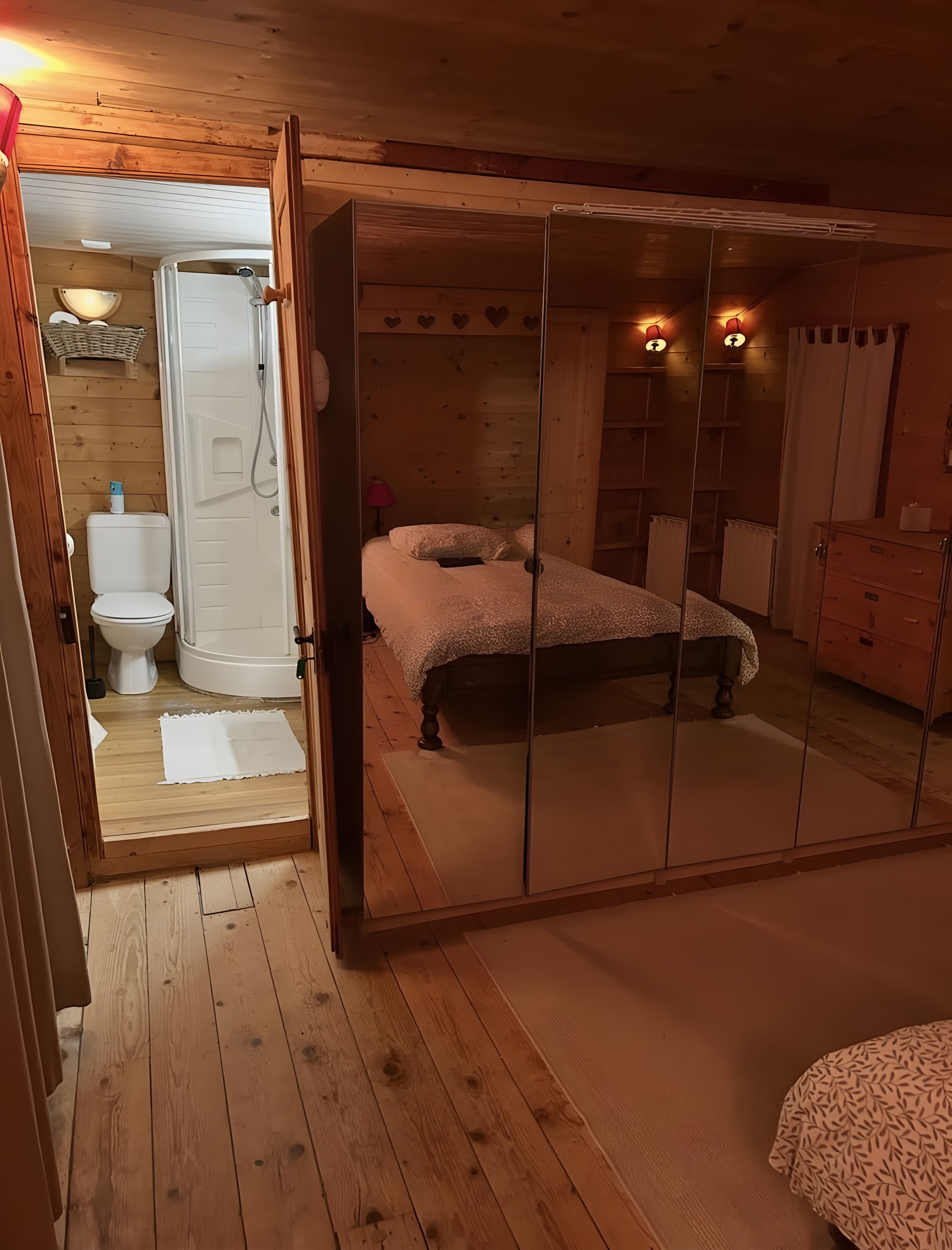 Una habitación con piso de madera, una cama con cubrecama blanco, un armario de madera y un baño con inodoro y ducha.