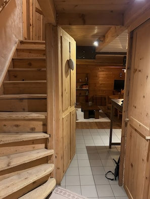 Un escalier en bois mène à une pièce où se trouvent une table à manger et des chaises, un téléviseur et une étagère remplie d’objets.