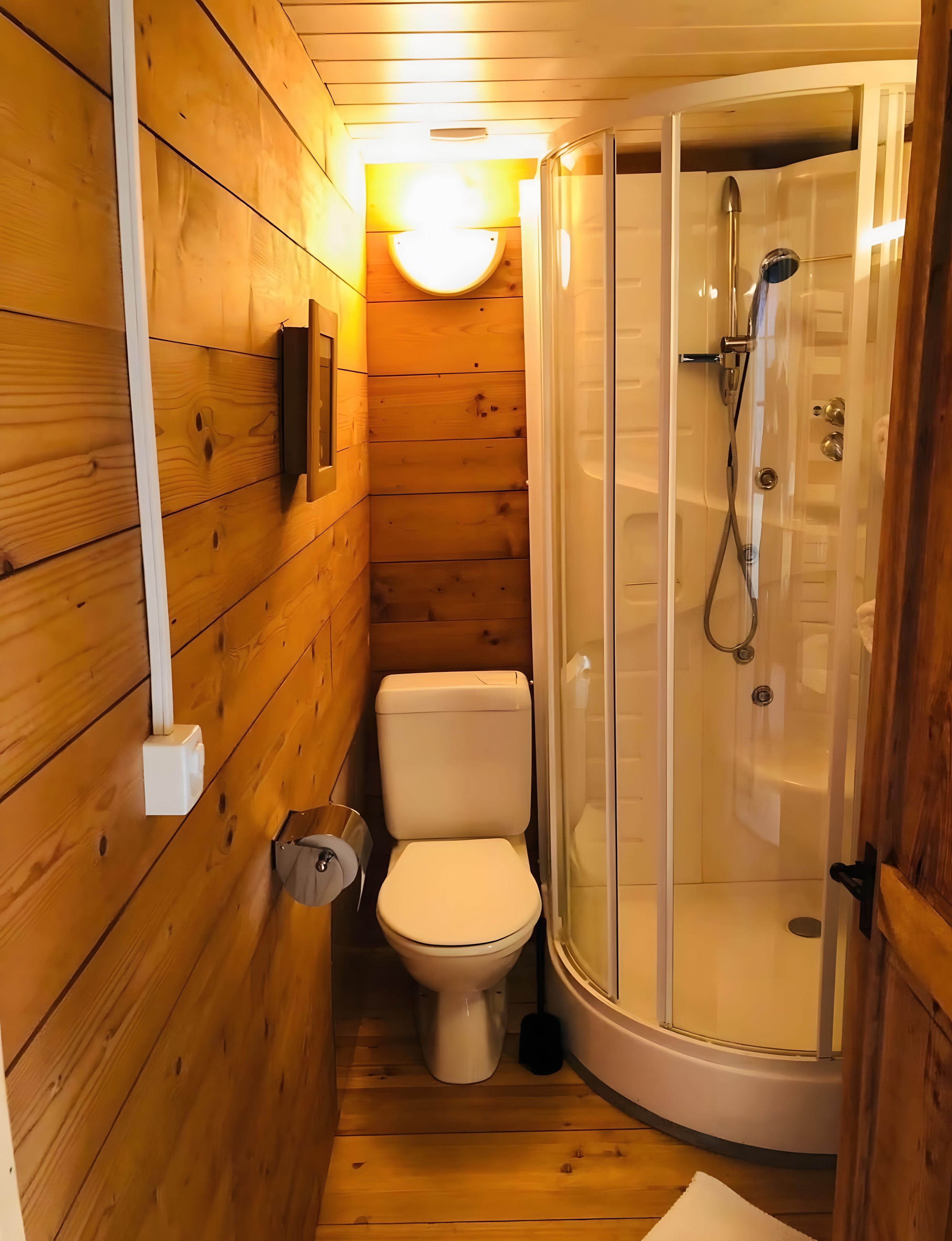 Une petite salle de bain en bois comprenant des toilettes, un espace douche et un luminaire fixé au mur.