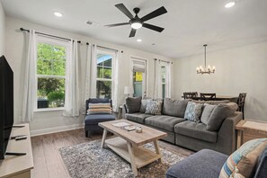 A living room with a ceiling fan, large windows with white curtains, a gray sofa with multiple cushions, a wooden coffee table, and a chandelier. - New on Program! Ready to Rent in The Townes at Barefoot #5206! Perfect Spot! (North Myrtle Beach)