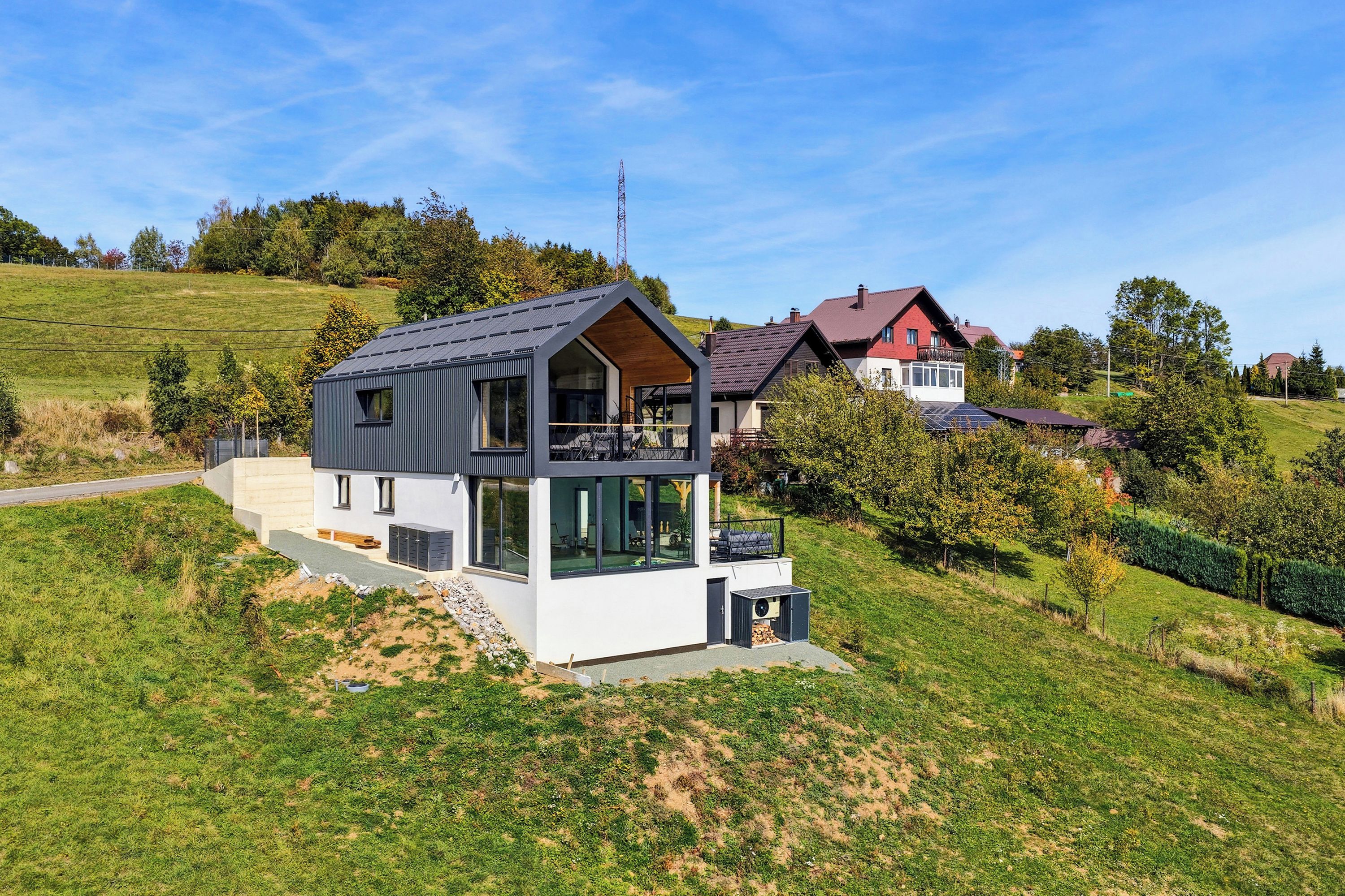 A modern two-story house with a balcony, situated on a grassy hillside.